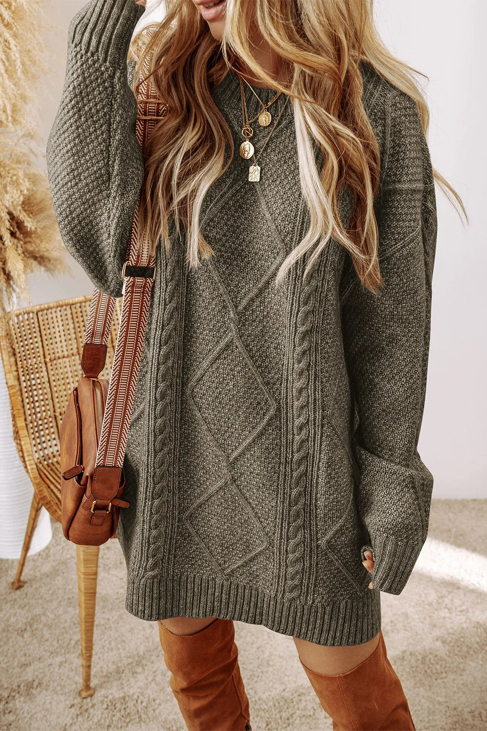 Sage cable-knit sweater dress with round neck and long sleeves, worn with layered gold necklaces and brown knee-high boots.