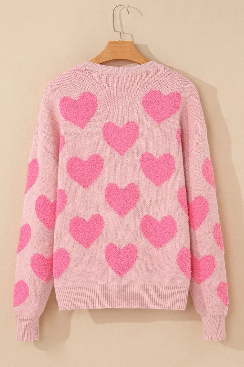 Blush pink sweater with bright pink heart jacquard pattern and round neck hanging on wooden hanger.