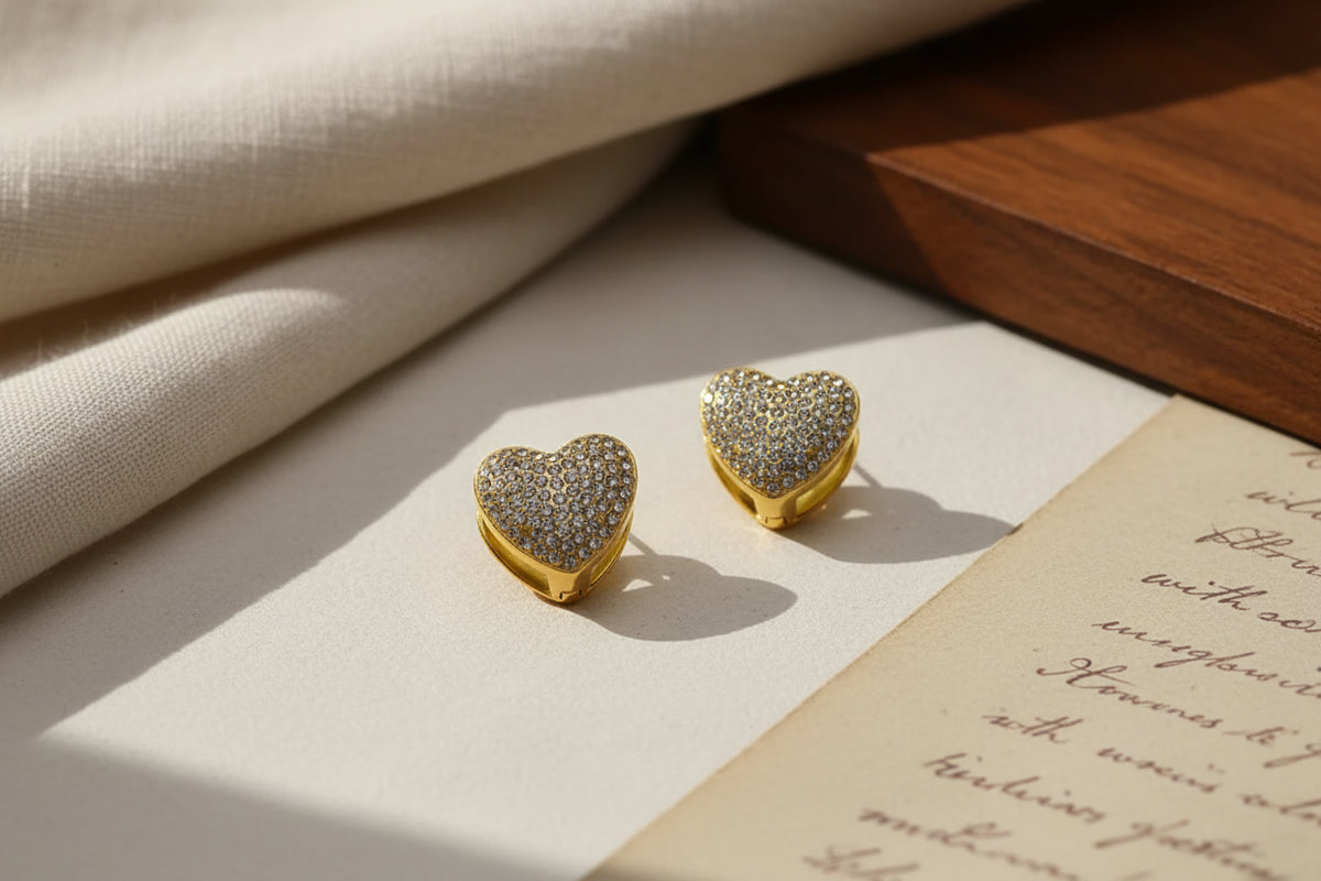 Gold heart-shaped stud earrings with pave diamond detailing on fabric and paper background.