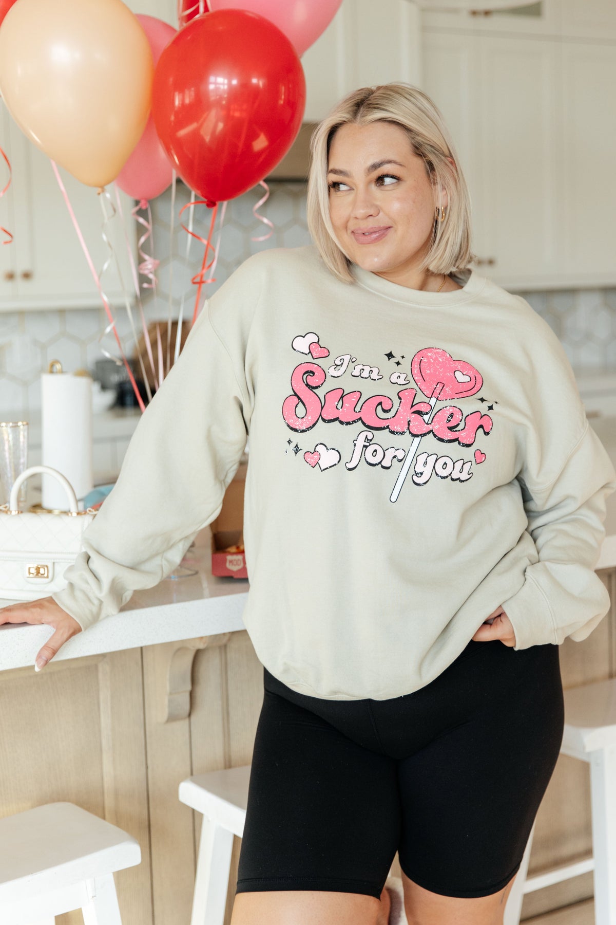 Light beige pullover sweatshirt with pink and white "I'm A Sucker For You" graphic and heart illustrations.
