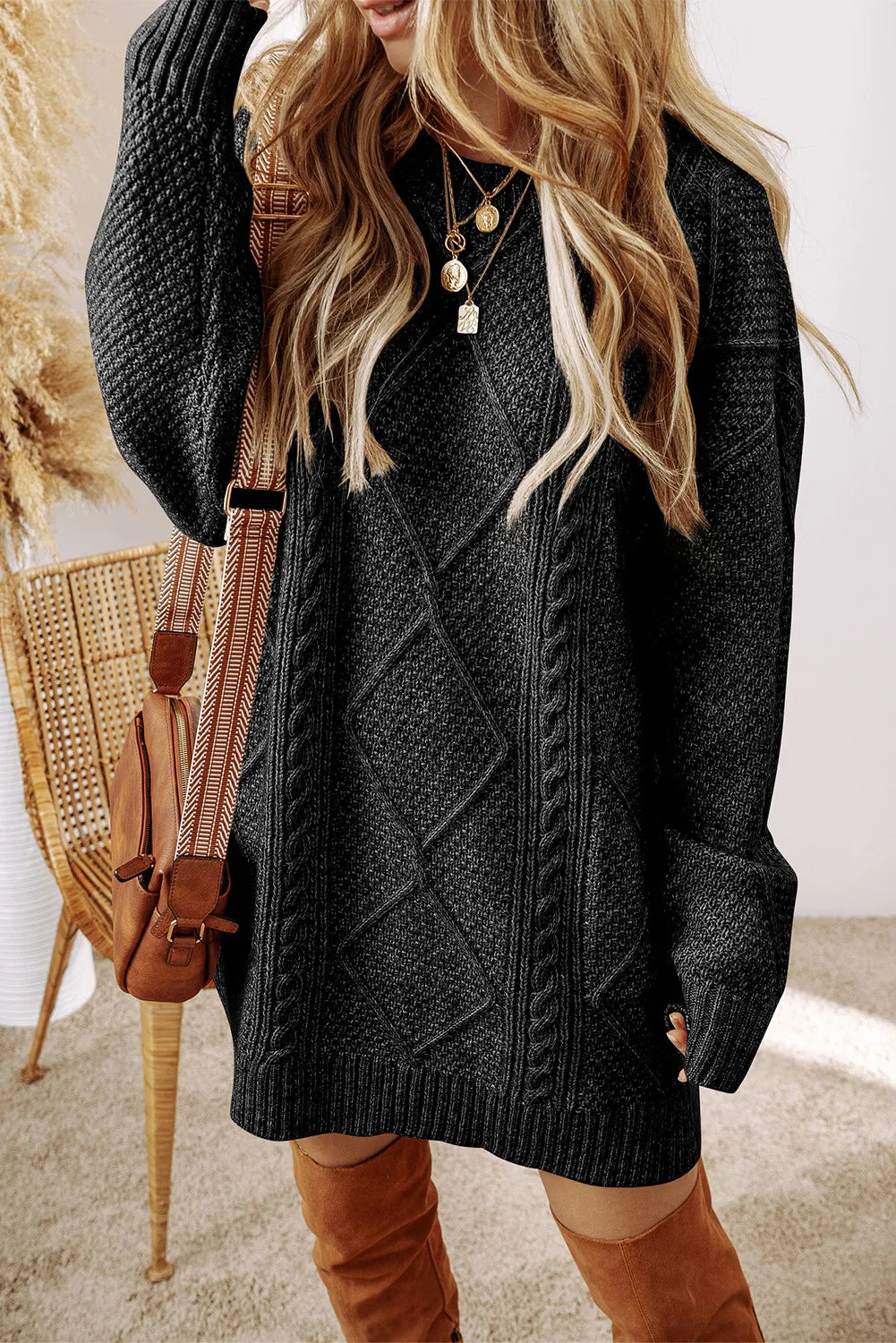 Black cable-knit sweater dress with round neck and long sleeves worn with layered gold necklaces and tan boots.