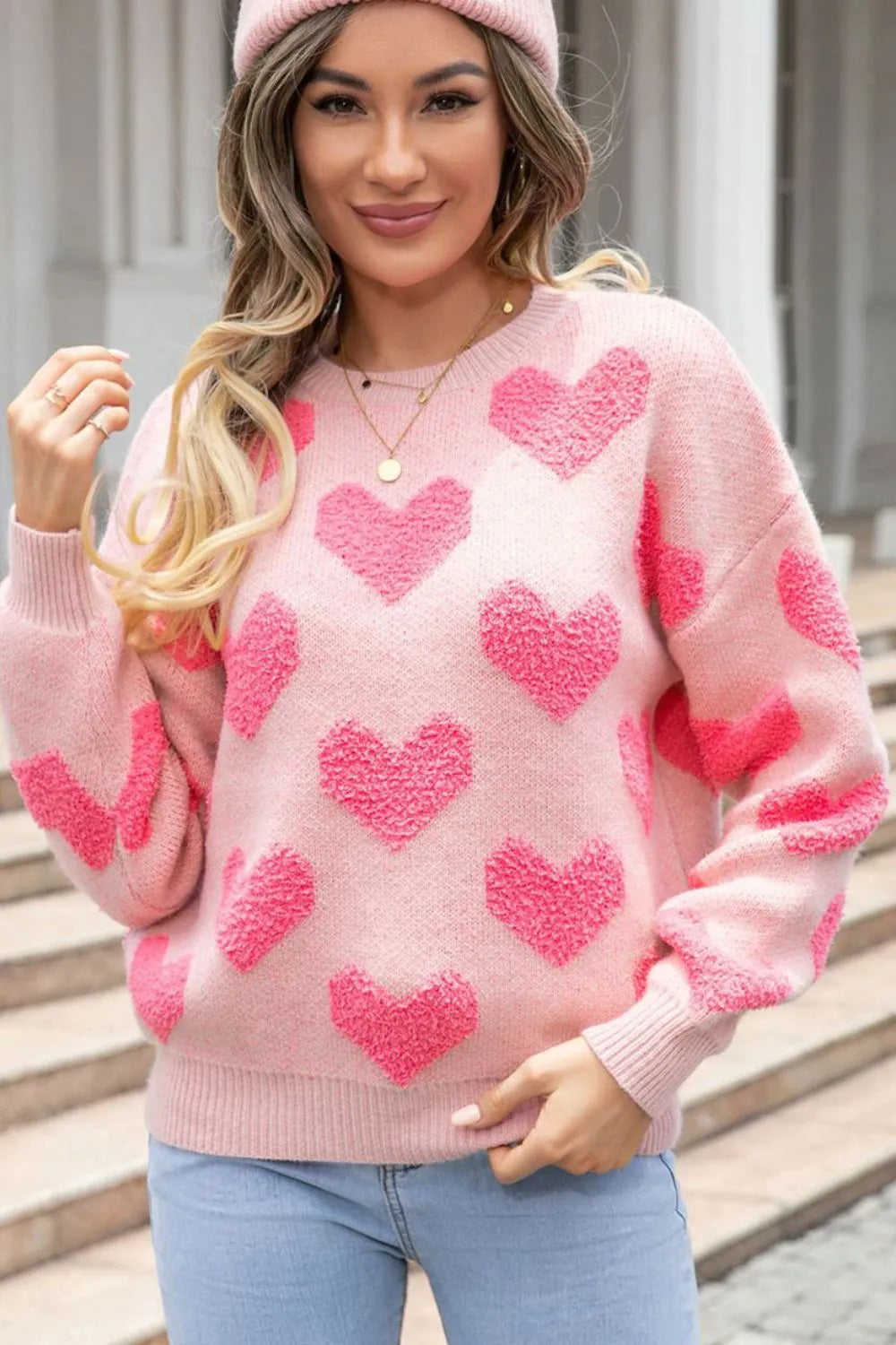 Blush pink sweater with large textured red hearts, round neck, and long sleeves, worn with matching pink hat.