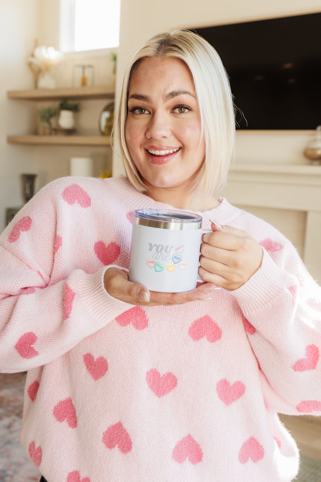 White double-walled travel mug with "You Are" multicolored text held by a woman in a pink sweater with red hearts.